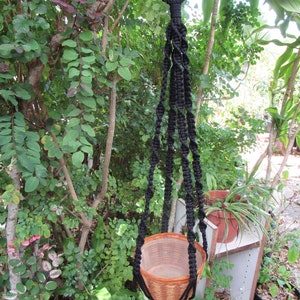 May include: Black macrame plant hanger with a woven basket hanging from it. The hanger is made of thick black cord and has a loop at the top for hanging.