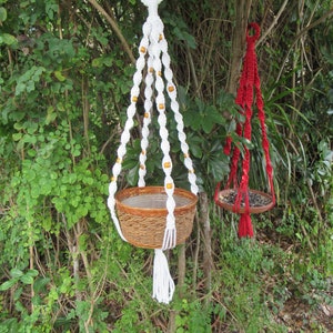 May include: Two macrame plant hangers with wooden pots. The hanger on the left is white with brown beads and the hanger on the right is red.