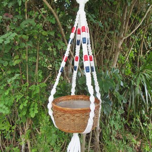 May include: White macrame plant hanger with red, white, and blue beads. The hanger is holding a brown woven pot.