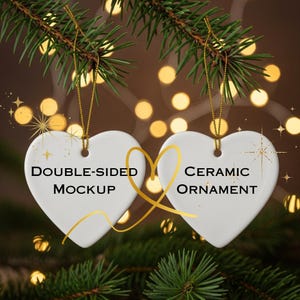 Puede incluir: Dos adornos de cerámica blancos en forma de corazón, colgados de una rama de pino verde. Cada adorno tiene un hilo dorado. El texto en el adorno izquierdo dice "DOUBLE-SIDED MOCKUP" y en el derecho "CERAMIC ORNAMENT".