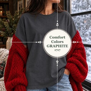 May include: A dark charcoal grey short-sleeve t-shirt featuring a round neckline. A circular graphic overlay in the centre displays the text Comfort Colors GRAPHITE 1717. The shirt is worn under a thick, chunky knit bright red cardigan sweater.