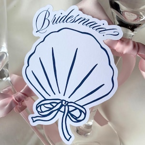 May include: A white seashell-shaped tag with the word "Bridesmaid?" in navy blue script. The tag has navy blue lines and a bow detail. A pink ribbon is tied around a champagne glass.