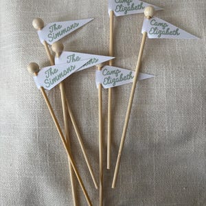 May include: A collection of six small pennant flags attached to thin wooden sticks with round wooden beads at the top. Three flags feature the green text The Simmons and three flags feature the green text Camp Elizabeth on white fabric.