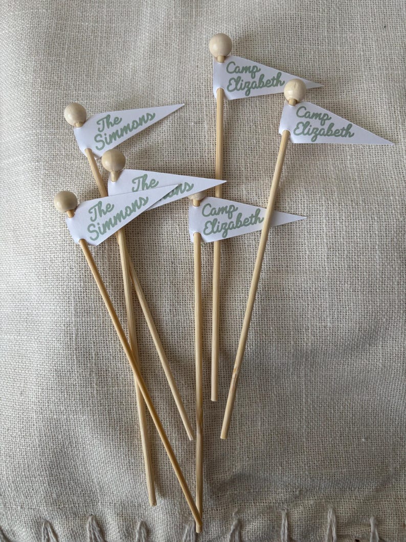 May include: A collection of six small pennant flags attached to thin wooden sticks with round wooden beads at the top. Three flags feature the green text The Simmons and three flags feature the green text Camp Elizabeth on white fabric.