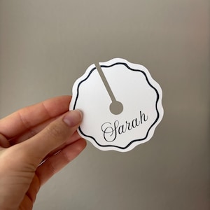 May include: A white, scalloped-edge wine glass charm with a black border and a slot for the glass stem. The charm has the name "Sarah" in elegant script. The charm is held in a hand.