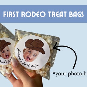 Peut inclure: Sacs transparents remplis de pop-corn, chacun avec un autocollant représentant un bébé portant un chapeau de cow-boy. L'autocollant indique "Andrew's first rodeo". Le texte "FIRST RODEO TREAT BAGS" est affiché sur fond bleu.
