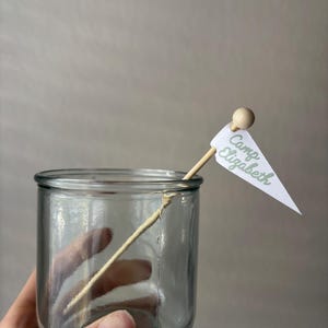 May include: A clear glass jar with a wooden stick inserted, featuring a small white triangular flag at the top. The flag is printed with the green text "Camp Elizabeth" and is topped with a small wooden bead.