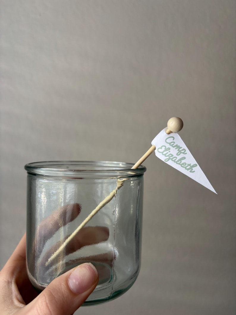 May include: A clear glass jar with a wooden stick inserted, featuring a small white triangular flag at the top. The flag is printed with the green text "Camp Elizabeth" and is topped with a small wooden bead.