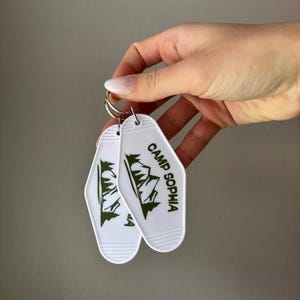 May include: Two white keychains with a retro motel key design. Each keychain features a green mountain graphic and the text "CAMP SOPHIA" in green. The keychains are held together by a silver metal ring.