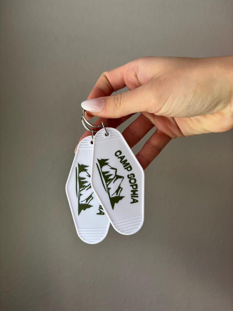 May include: Two white keychains with a retro motel key design. Each keychain features a green mountain graphic and the text "CAMP SOPHIA" in green. The keychains are held together by a silver metal ring.