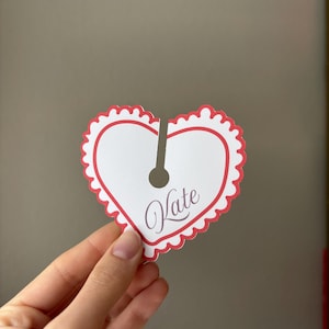 May include: A heart-shaped tag with a scalloped red border and the name "Kate" in cursive. The tag is white with a vertical slit and a hole at the top. The tag is held by a hand.