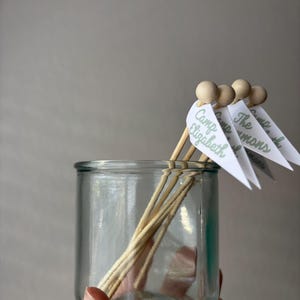 May include: A clear glass jar containing several wooden skewers, each topped with a small wooden bead and a white triangular paper flag. The flags feature green printed text that reads Camp Elizabeth, The Simmons, and Camp.