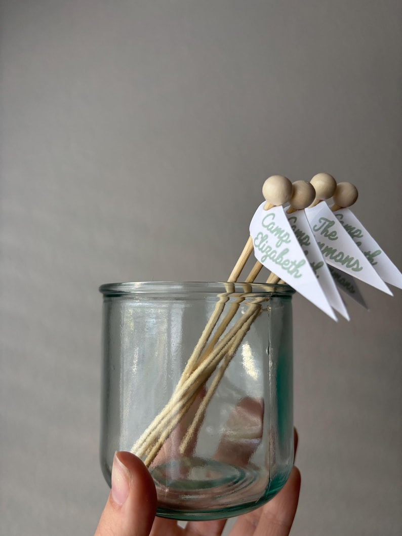 May include: A clear glass jar containing several wooden skewers, each topped with a small wooden bead and a white triangular paper flag. The flags feature green printed text that reads Camp Elizabeth, The Simmons, and Camp.