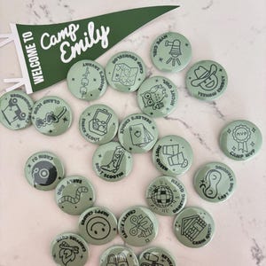 May include: A selection of sage green camp-themed badges featuring illustrations and text, such as a cowboy hat, a disco ball, and the words "Camp Emily." A green pennant banner that says "Welcome to Camp Emily" is also included.