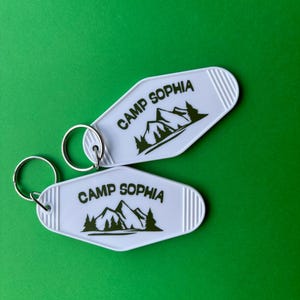 May include: Two white keychains with the text "CAMP SOPHIA" and a mountain and tree graphic in black. The keychains have a retro motel style with a silver key ring and are set against a green background.