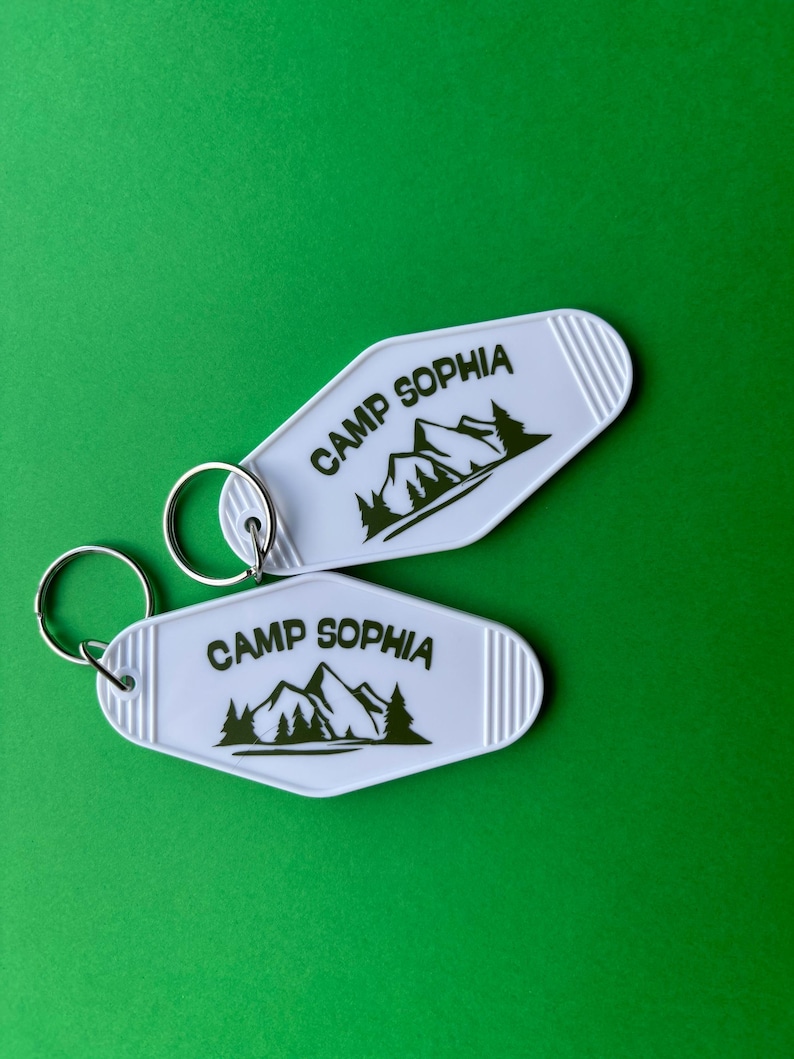 May include: Two white keychains with the text "CAMP SOPHIA" and a mountain and tree graphic in black. The keychains have a retro motel style with a silver key ring and are set against a green background.