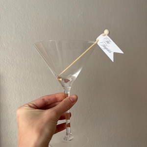 May include: A clear glass martini glass with a long stem and a wide, conical bowl. Inside the glass rests a wooden cocktail pick topped with a small round bead and a white paper flag featuring the text The Hayeses in a black script font.