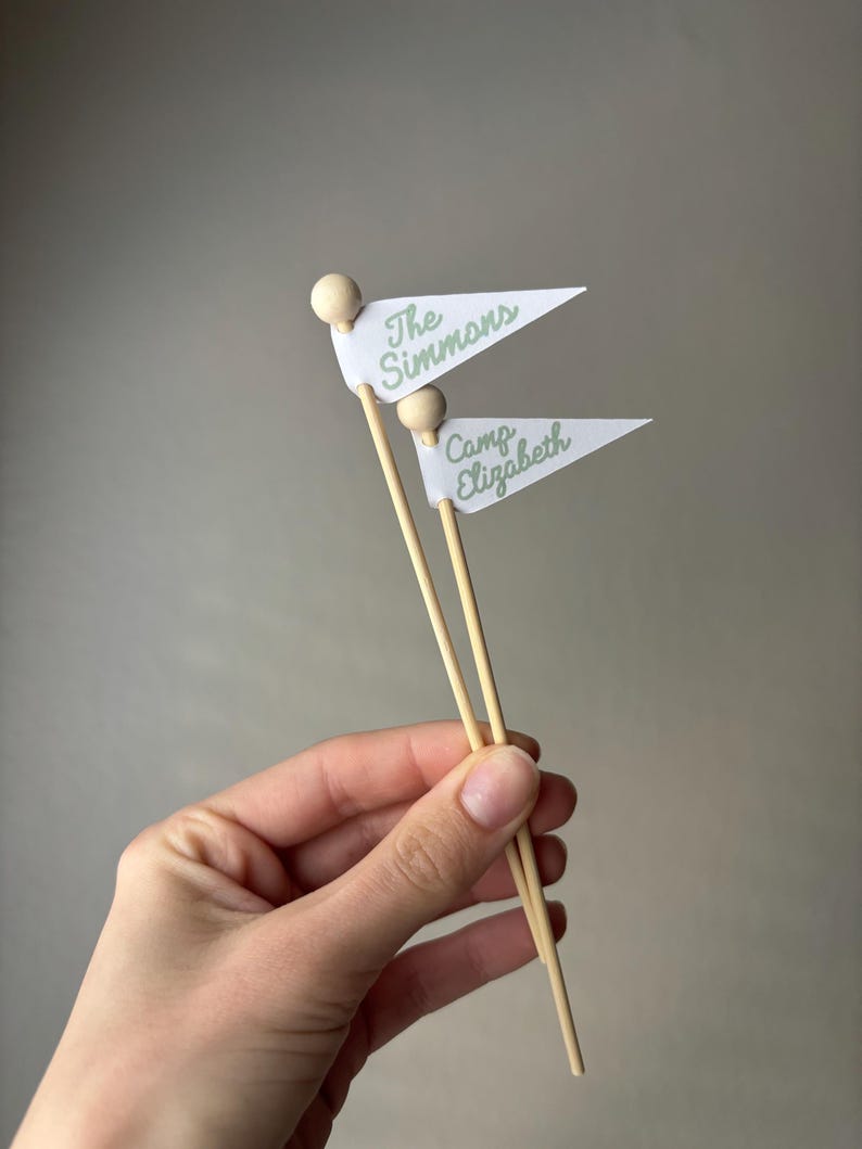 May include: Two small white triangular pennants attached to thin wooden sticks with round wooden beads at the top. The top pennant features the green text The Simmons, and the bottom pennant features the green text Camp Elizabeth.