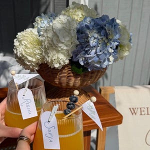 May include: Two ribbed glass tumblers filled with orange juice, each featuring a white tag clipped to the rim with the names Regina and Sofia, and a small white flag labelled Baby Castillo Lugo, alongside a wicker basket of blue and white hydrangeas.