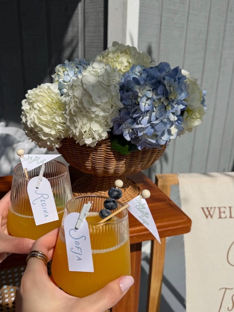 May include: Two ribbed glass tumblers filled with orange juice, each featuring a white tag clipped to the rim with the names Regina and Sofia, and a small white flag labelled Baby Castillo Lugo, alongside a wicker basket of blue and white hydrangeas.