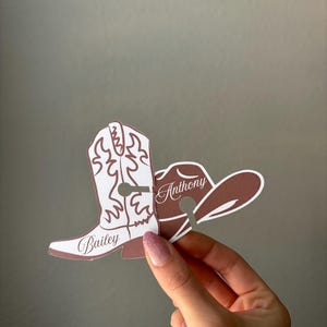 May include: A brown and white cowboy boot and hat cutout with the names "Bailey" and "Anthony" printed on them. The boot has decorative stitching and the hat is a classic cowboy style. The cutouts are held up by a hand.