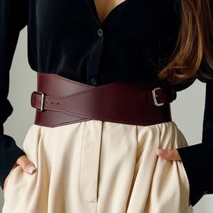 May include: A wide, burgundy leather belt with a rectangular buckle and a matching leather strap. The belt is worn over a black button-down shirt and beige pants. The belt is a fashion accessory.