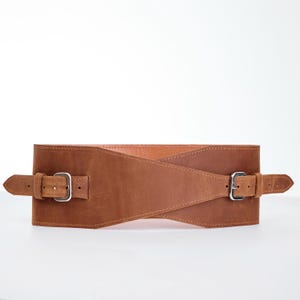 May include: A wide, brown leather belt with two silver-colored buckles. The belt has a crisscross design in the front and is designed to cinch the waist. The leather has a distressed look.