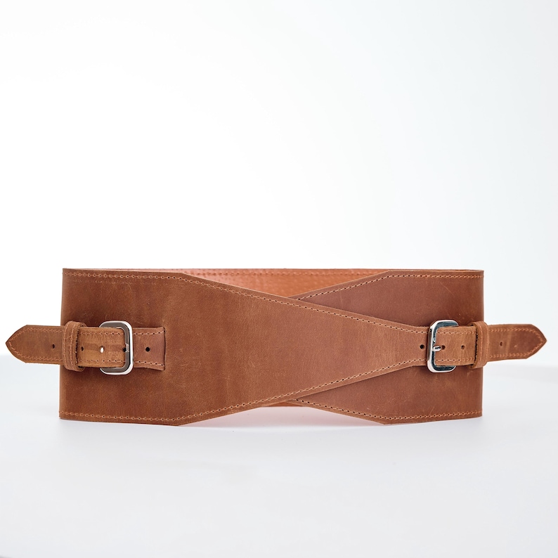 May include: A wide, brown leather belt with two silver-colored buckles. The belt has a crisscross design in the front and is designed to cinch the waist. The leather has a distressed look.