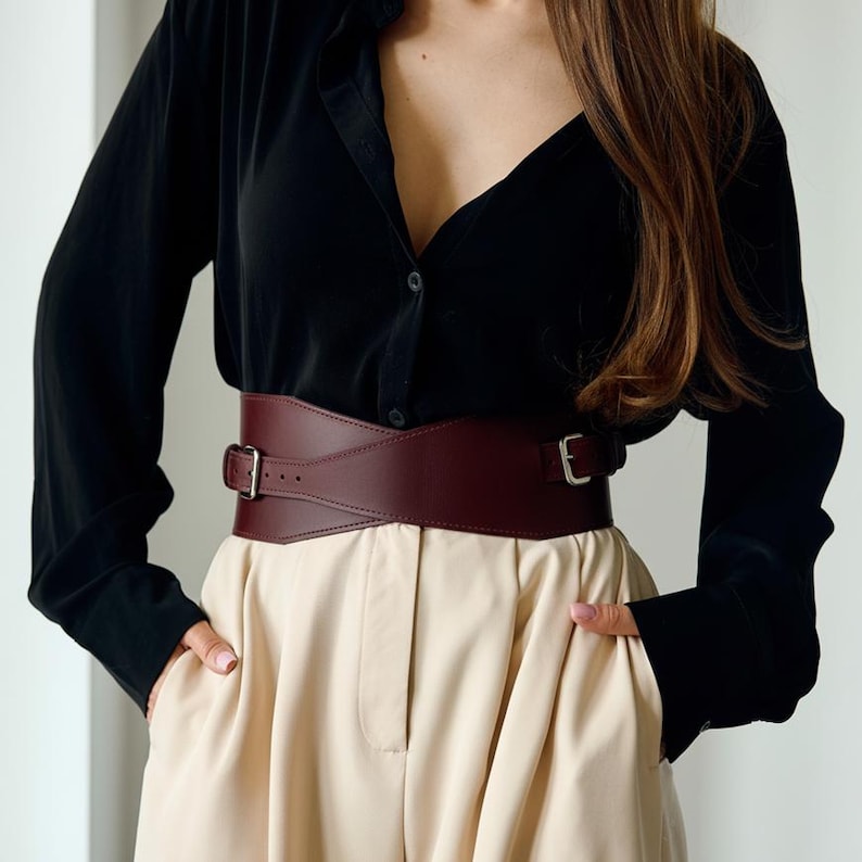 May include: A wide, burgundy leather belt with a rectangular buckle and a matching leather strap. The belt is worn over a black button-down shirt and beige pants. The belt is a fashion accessory.