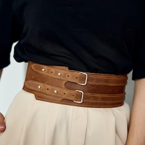 Puede incluir: Cinturón ancho de cuero marrón con dos filas de hebillas y herrajes plateados. El cinturón tiene un aspecto texturizado y se asegura con correas ajustables. El diseño es de estilo corsé, adecuado para ceñir la cintura.