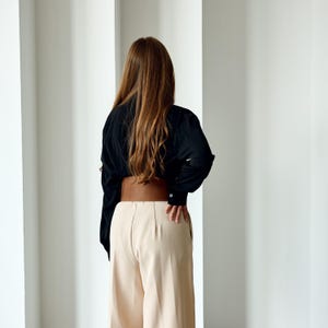 May include: A person wearing a black long-sleeve shirt, tan pants, and a wide brown belt. The person is facing away from the camera, with long brown hair. The pants are high-waisted and wide-legged.