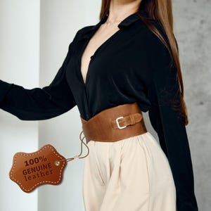 May include: A wide, brown leather belt with a silver buckle. The belt is cinched at the waist and has a tag that reads "100% GENUINE leather". The belt is worn over a black shirt and tan pants.