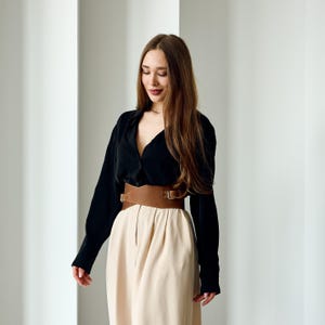 May include: A woman wearing a black long-sleeve shirt, a wide brown leather belt with a buckle, and beige pants. The outfit is styled in front of a white background with vertical lines.