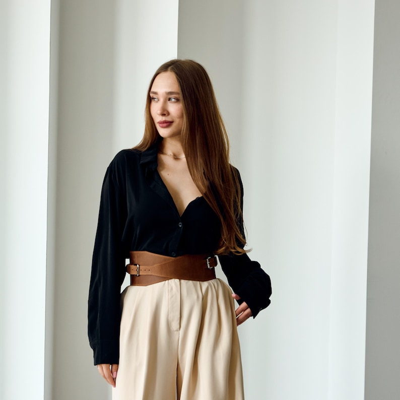 May include: A black button-down shirt paired with beige pants and a wide brown leather belt. The belt has a double-strap design with buckle closures. The outfit is styled in a minimalist aesthetic.