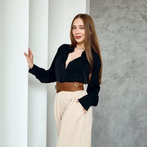 May include: A woman wearing a black long-sleeve shirt, tan pants, and a wide brown belt with a gold buckle. The outfit is styled with a neutral color palette. The background features white pillars and a gray wall.