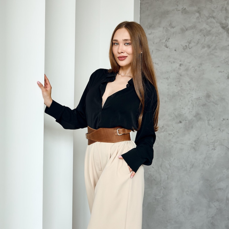 May include: A woman wearing a black long-sleeve shirt, tan pants, and a wide brown belt with a gold buckle. The outfit is styled with a neutral color palette. The background features white pillars and a gray wall.