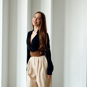 May include: A black long-sleeve shirt with a deep V-neck, paired with high-waisted, beige pants. A wide brown belt with a rectangular buckle cinches the waist. The outfit is styled in a modern, minimalist aesthetic.