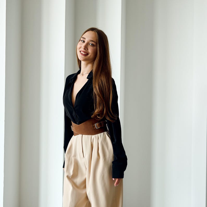 May include: A black long-sleeve shirt with a deep V-neck, paired with high-waisted, beige pants. A wide brown belt with a rectangular buckle cinches the waist. The outfit is styled in a modern, minimalist aesthetic.