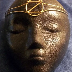 May include: A gold-toned headband with a geometric design featuring a circle with a Z inside. The headband is on a black mannequin head.