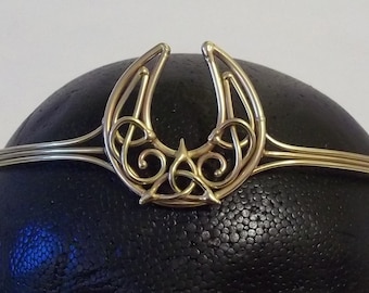 May include: A gold-colored metal tiara with a horseshoe-shaped design. The tiara features a central Celtic knot design and is displayed on a black surface.