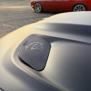 May include: Close-up of a black car hood with a carbon fiber fuel door featuring a silver Hellcat logo. A red Dodge Challenger is visible in the background, parked in a lot during the golden hour.