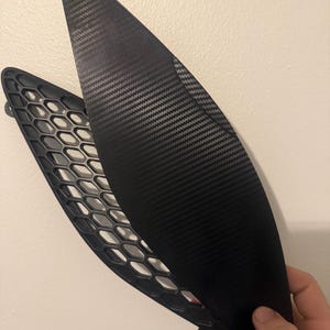 May include: Two black automotive parts. One is a curved, carbon fiber textured panel. The other is a black plastic honeycomb-patterned grill. The parts are likely for a vehicle's exterior.