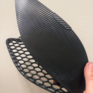 May include: A black carbon fiber car part with a honeycomb-patterned mesh. The part has a curved, aerodynamic shape and a textured surface, likely for improved performance and aesthetics.