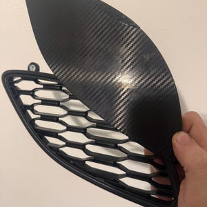 May include: A black car part with a carbon fiber texture, held up to show the detail. The part has a honeycomb-patterned grill. The carbon fiber piece is curved and has a glossy finish.