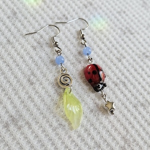 May include: A pair of handmade earrings with silver-tone hooks. One earring features a red ladybug charm with black spots, while the other has a green leaf-shaped charm. Both earrings have blue beads and a silver spiral design.