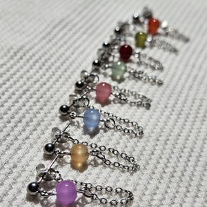 May include: A collection of colorful beaded earrings. Each earring features a small, round bead in a different color, including purple, yellow, blue, pink, green, red, and orange. The beads are connected to a chain and a silver post.
