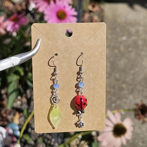May include: A pair of handmade earrings displayed on a brown card. One earring features a red ladybug charm, while the other has a green leaf and silver spiral. Both have blue beads and silver hooks. The background includes pink flowers.