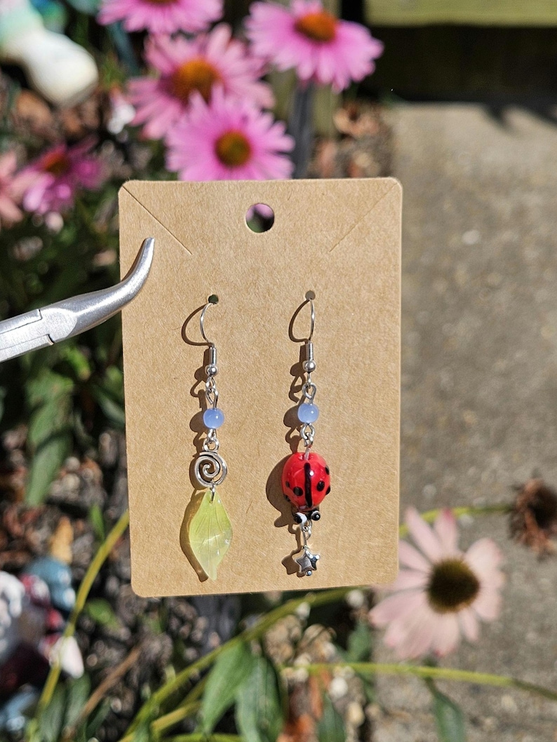 May include: A pair of handmade earrings displayed on a brown card. One earring features a red ladybug charm, while the other has a green leaf and silver spiral. Both have blue beads and silver hooks. The background includes pink flowers.