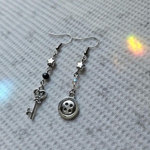 May include: A pair of silver-toned dangle earrings. One earring features a key charm and black beads, while the other has a button charm. Both earrings have star-shaped accents and are suspended from a hook.