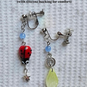 May include: Clip-on earrings with stainless steel adjustable clips. One earring features a red ladybug charm with black spots and a star pendant. The other earring has a green leaf charm and a spiral design. Both have blue beads.